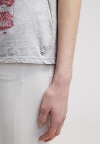 Grey cotton T-shirt featuring a red graphic print, with a small star accent at the hem and a relaxed fit paired with white pants.