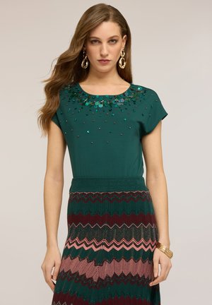 Frau mit langem, welligem Haar trägt ein grünes, verziertes Oberteil, einen mehrfarbigen Rock mit Zickzackmuster, goldene Ohrringe und mehrere goldene Armbänder am Handgelenk.