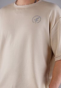 Beige short-sleeved t-shirt with a crew neck, featuring a textured finish, stitched hem, and a small gray logo on the left chest.