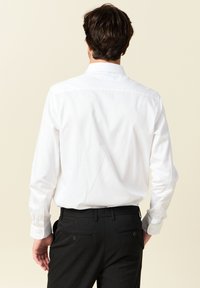Camisa blanca de manga larga con cuello abotonado, combinada con pantalones de vestir negros que tienen dos bolsillos traseros y un acabado texturizado.