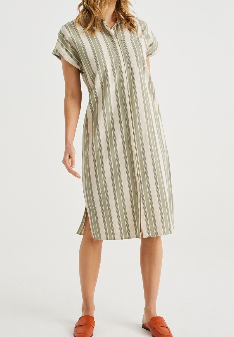 Robe en lin rayée beigne et verte avec des manches courtes, une poche frontale et des fentes latérales, associée à des chaussures orange à enfiler.