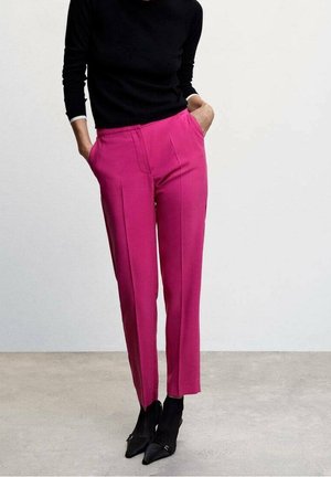 Person wearing fitted black long-sleeve top tucked into high-waisted bright pink trousers with hands in pockets and black pointed ankle boots.