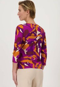 Paarse top met abstracte oranje en bordeaux bladpatronen. Driekwart mouwen en een ronde halslijn. Materiaal lijkt soepel en rekbaar.