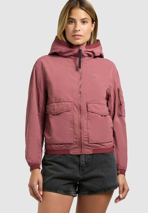 Nő poros rózsaszín kapucnis dzsekiben, nagy elülső zsebekkel és fekete farmerrövidnadrágban, világosszürke egyszínű háttér előtt állva.