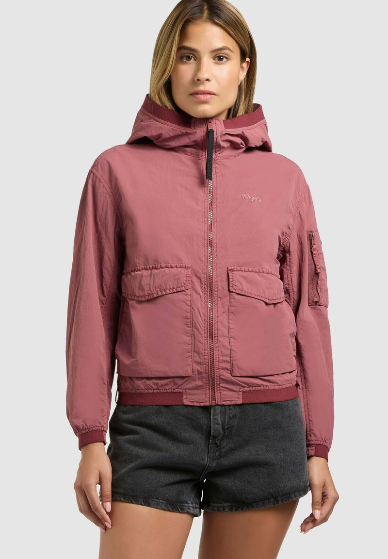 Donna che indossa una giacca con cappuccio color rosa polvere con grandi tasche frontali e pantaloncini di denim nero, in piedi davanti a uno sfondo grigio chiaro uniforme.