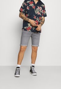 Osoba ubrana w granatową koszulę w kwiaty z krótkim rękawem, szare krótkie spodenki z podwiniętymi nogawkami, czarne trampki za kostkę i białe skarpetki, stojąca na jednolitym tle.