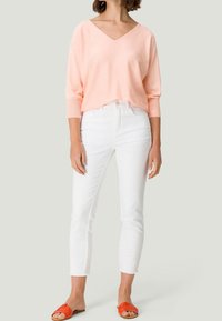 Femme portant un pull V-neck pêche, un jean blanc slim-fit crop et des sandales à glisser orange, se tenant devant un fond gris clair.