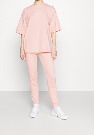 Persoon draagt een loszittend lichtroze T-shirt met korte mouwen en bijpassende roze joggerbroek met witte sneakers.