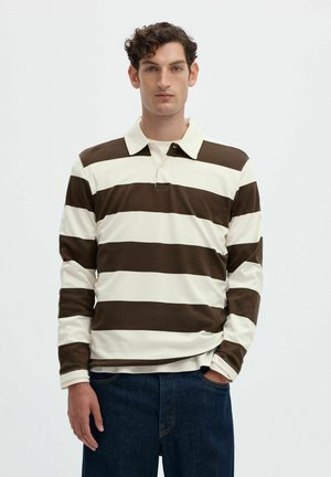 Homme aux cheveux bouclés foncés portant un maillot de rugby rayé marron et blanc à manches longues et un jean bleu foncé, debout avec une main dans la poche.