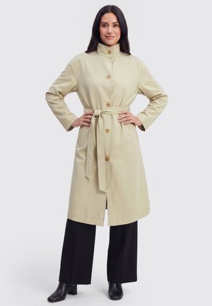 Kvinde iført en beige trenchcoat med bælte, sorte vidde bukser og sorte ankelstøvler, der står med hænderne på hofterne.
