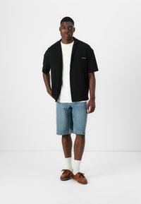 Schwarzes kurzärmeliges Shirt über einem weißen T-Shirt, kombiniert mit hellblauen Denim-Shorts und braunen Bootsschuhen. Strukturiertes Material, lässiges Design.
