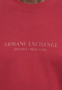 Raudona medvilninė megztinis su apvalia iškirpte, su reljefiniu tekstu "ARMANI EXCHANGE MILANO / NEW YORK" priekyje, turintis lygią tekstūrą.