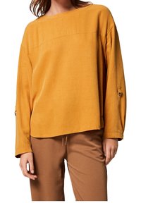 Top lungo a maniche con scollo a barca di colore giallo senape. Caratterizzato da una vestibilità ampia, polsini con dettagli a bottone e un tessuto morbido e testurizzato.
