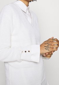 Pull blanc en tricot avec un col en V, superposé à une chemise blanche. Le pull présente des poignets côtelés ornés de boutons décoratifs dorés.