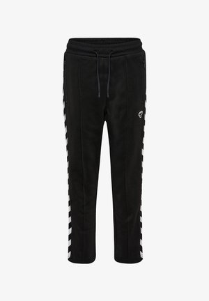 Dziecko o kręconych włosach ubrane w czarny dres z białymi pasami chevron i różowymi trampkami, stojące na jednolitym tle.