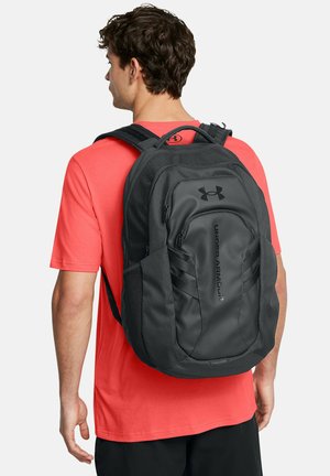 Black backpack with a sleek design, featuring a front pocket, textured materials, and mesh side pockets, worn on a red shirt.