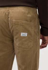 Pantalons en velours côtelé beige avec une texture à côtes verticales, comportant une poche arrière et une étiquette. Associés à un haut texturé brun foncé.