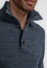 Suéter de punto azul oscuro con un cuello alto, que presenta una cremallera y dos grandes botones. La tela tiene un patrón texturizado.