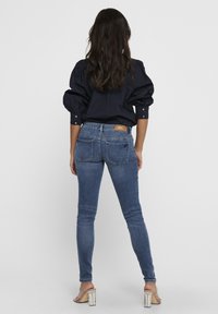 Camisa de manga abullonada en azul oscuro combinada con jeans ajustados de algodón azul claro. Características notables incluyen bolsillos y una etiqueta de marca en la cintura.