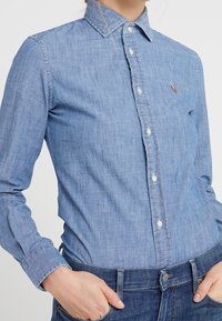 Camisa de mezclilla azul claro con collar abotonado, costuras visibles y un pequeño logo en el lado izquierdo del pecho. Mangas largas con puños abotonados.