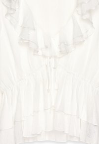 Blusa blanca de tela texturizada, con volantes en capas a lo largo del escote y el dobladillo, y un cierre con corbata en el frente.