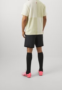 T-shirt à rayures en jaune clair et crème, shorts noirs, chaussettes noires et chaussures de soccer roses avec une marque.