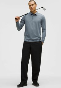 Světle modrá polokošile s dlouhým rukávem a límečkem, v kombinaci s černými kalhotami a černými teniskami; drží golfovou hůl.