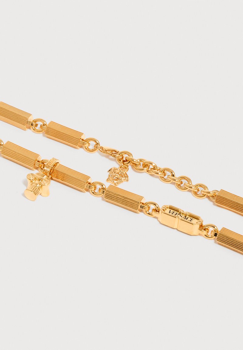 Collier en chaîne dorée avec maillons rectangulaires, pendentif tête de Méduse, pendentif chapiteau de colonne et fermoir gravé Versace sur fond blanc.