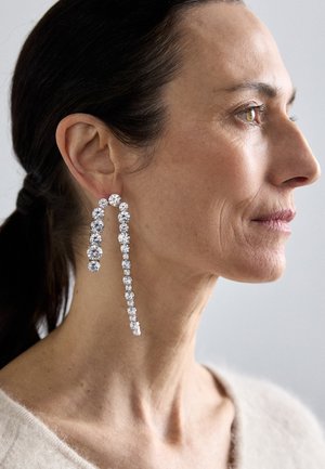 De longues boucles d'oreilles pendantes en cristaux clairs étincelants, avec un design linéaire de tailles graduées, portées sur un teint clair et lisse.