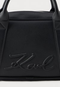 Melnas ādas rokas soma ar izšūtām rokturiem un uzspiedušo parakstu "Karl" uz priekšējā paneļa, kā arī redzamu rāvējslēdzēju augšpusē.