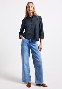 Camicia nera button-up con dettagli ricamati sull'orlo, abbinata a jeans a gamba larga di colore blu chiaro e sandali marroni.