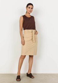 Jupe en coton beige avec une ceinture nouée, des poches latérales et une coupe ajustée, accompagnée d’un haut sans manches marron et de sandales à talons blocs marron foncé.