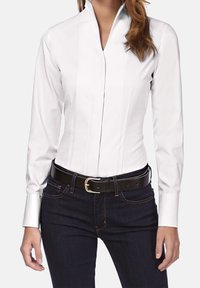 Camicia bianca a maniche lunghe con colletto alla cinese, caratterizzata da un design aderente e sottili cuciture verticali, indossata con jeans in denim scuro e una cintura nera.