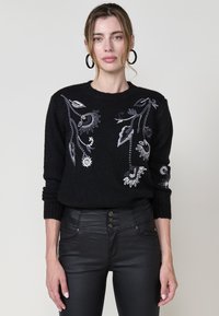 Donna che indossa un maglione nero con ricami floreali grigi e pantaloni in pelle neri a vita alta, rivolta in avanti con espressione neutra.