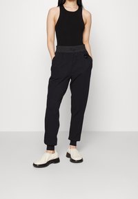 Svart ärmlös topp i kombination med svarta tapered joggers med elastisk midja och en sidologopatch, kompletterat med vita slip-on skor.