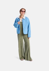 Prošívaná modrá bunda s bílým límcem, nošená přes zelený kordurový overal. Sluneční brýle a jednoduchý náhrdelník doplňují outfit.