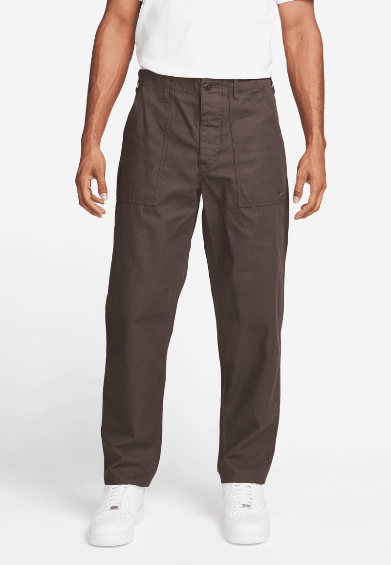 Ruskeat, kevyet cargo-housut, joissa on löysä malli, useita etupusseja ja napitus, yhdistettynä valkoisiin tennareihin ja t-paitaan.