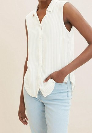 Kobieta o średnio-ciemnej karnacji, ubrana w białą, bezrękawnikową koszulę na guziki i jasnoniebieskie jeansy, z ręką w kieszeni.