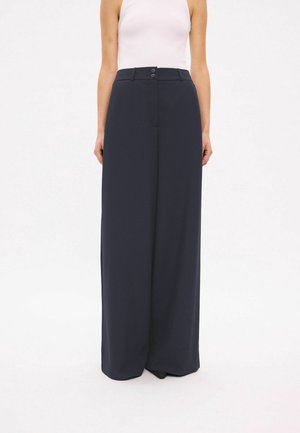 Femme portant un pantalon taille haute à jambes larges bleu marine foncé et un haut blanc sans manches sur fond blanc uni.