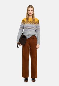 Szary dziergany sweter z żółtymi, fioletowymi i brązowymi wzorami, w zestawieniu z brązowymi spodniami o szerokich nogawkach. Modelka trzyma ciemnobrązową torbę.