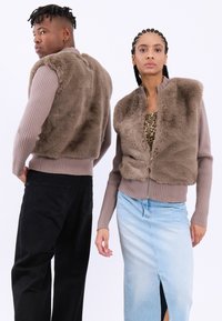 Vesta din blană sintetică maro cu mâneci lungi cu textură canală; modelul din stânga poartă pantaloni negri, iar cel din dreapta poartă o fustă din denim deschisă cu o deschidere frontală.