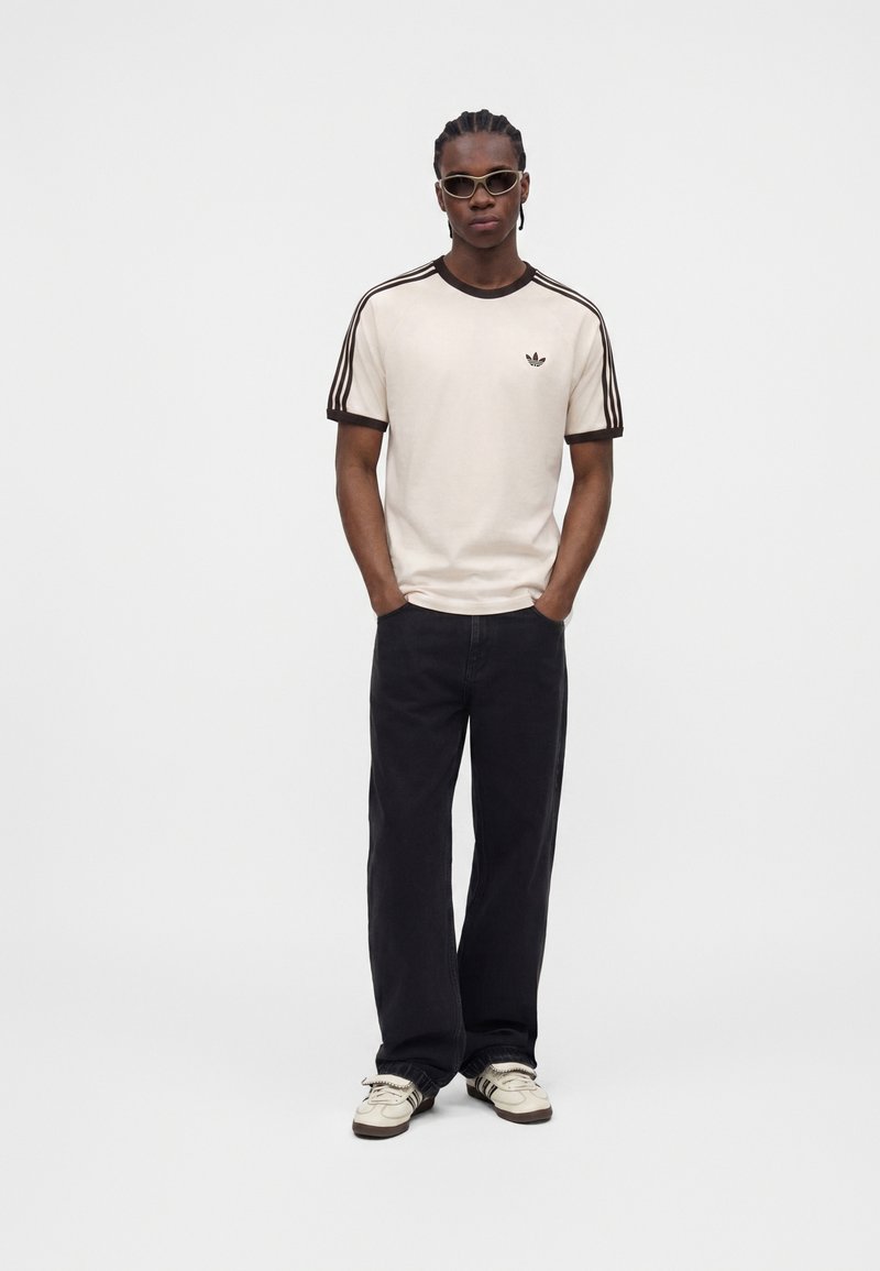Mann mit geflochtenen Haaren, der ein beiges Adidas-T-Shirt mit schwarzen Streifen, schwarze weite Hosen, weiße Turnschuhe und eine Sonnenbrille trägt und mit den Händen in den Taschen steht.