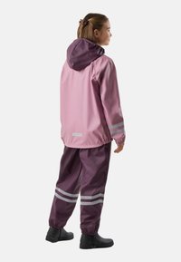Fille portant une veste de pluie rose et violette et un pantalon avec des bandes réfléchissantes, debout et regardant vers la droite sur fond blanc.