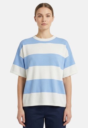 Donna con pelle chiara e capelli castani che indossa una maglietta a righe orizzontali bianche e azzurro chiaro a maniche corte e pantaloni scuri.