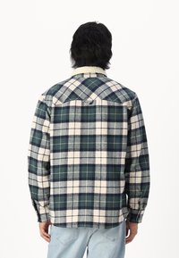 Chemise en flanelle avec un motif à carreaux bleu marine, vert et crème. Présente un col en polaire crème et des poignets boutonnés. Détail du dos visible.