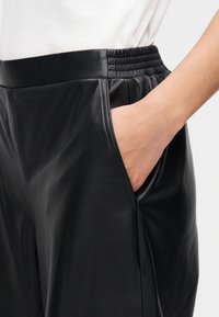 Calças de couro pretas com cintura elástica, apresentando bolsos laterais e textura lisa, exibindo um caimento ajustado e design simples.