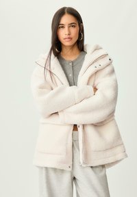 Veste en fausse fourrure rose clair, avec un zip à l'avant, une capuche ajustable et deux poches latérales. Texture douce avec une coupe décontractée.