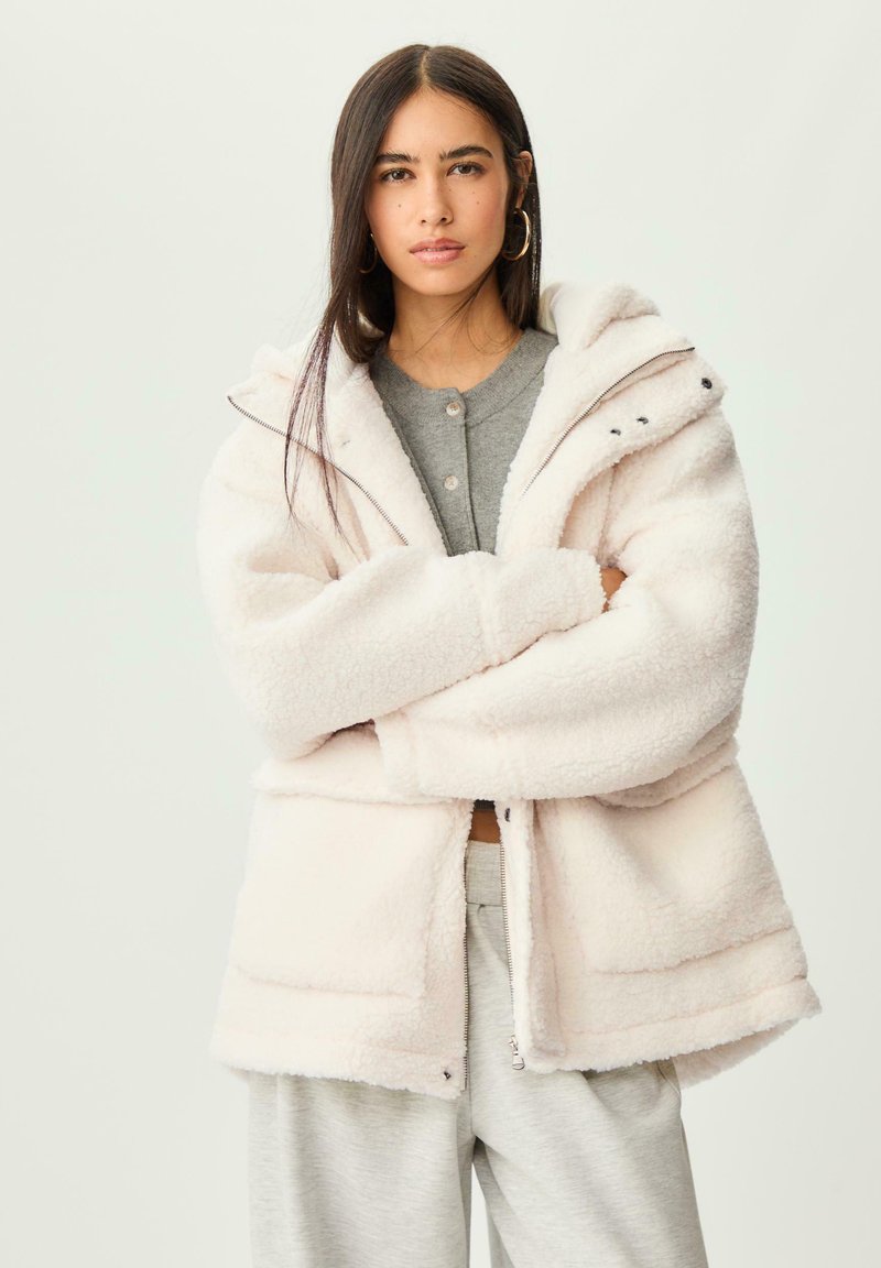 Veste en fausse fourrure rose clair, avec un zip à l'avant, une capuche ajustable et deux poches latérales. Texture douce avec une coupe décontractée.