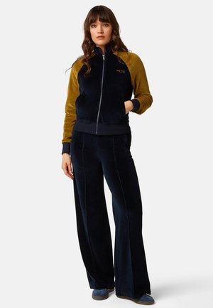 Mujer de pie con chaqueta con cremallera en azul marino y mostaza, pantalones anchos azul marino a juego y zapatos azules sobre fondo blanco.