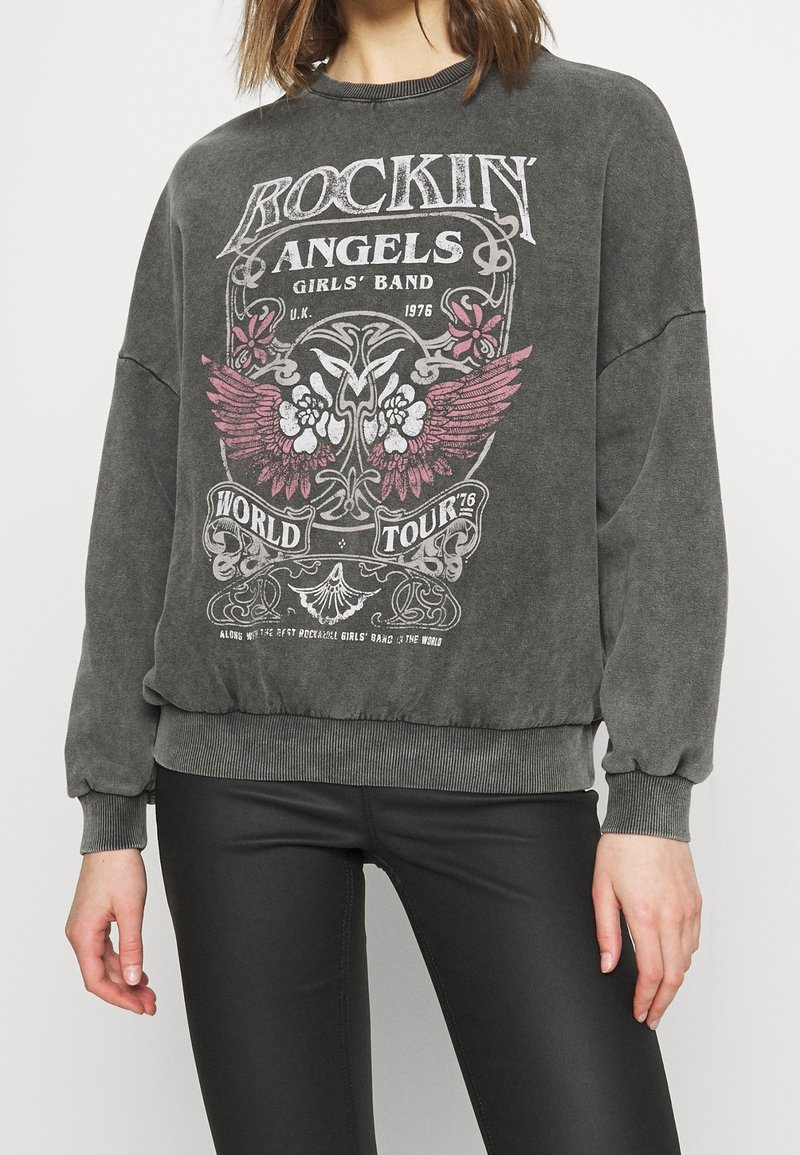 Woman wearing faded charcoal sweatshirt with "Rockin' Angels Girls' Band World Tour" text and winged floral graphic, paired with black leggings.
